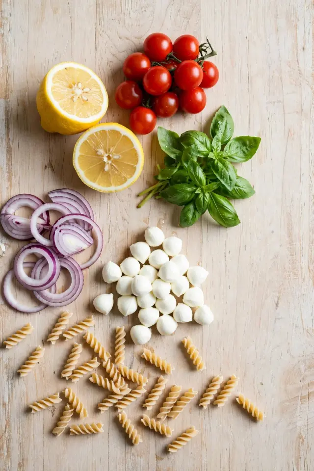 Lemon Basil Pasta Salad: The Bright, Make-Ahead Side Everyone Asks For 1 Fresh ingredients for lemon basil pasta salad including lemons, basil, cherry tomatoes, mozzarella pearls, and dried pasta laid out on a wood surface